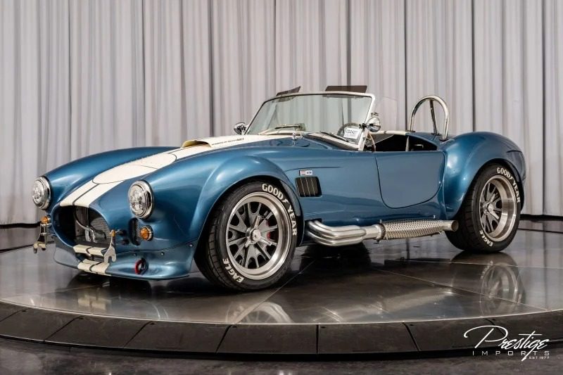 A blue Shelby Cobra sports car with white racing stripes, chrome side pipes, and Goodyear tires is displayed on a rotating platform among classic muscle cars in a showroom.