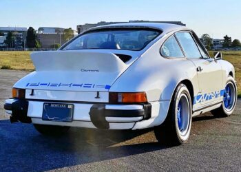 A white classic 1974 Carrera 2.7 MFI for sale, with blue accents and a Montana license plate, is parked on an open lot with grass and buildings in the background—this legend can outrun a Porsche 911 Turbo 3.0.