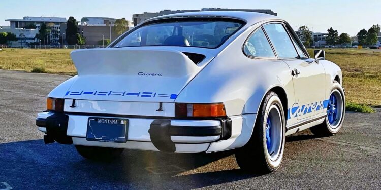 A white classic 1974 Carrera 2.7 MFI for sale, with blue accents and a Montana license plate, is parked on an open lot with grass and buildings in the background—this legend can outrun a Porsche 911 Turbo 3.0.