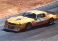 Yellow race car with number 15 and "YARBOROUGH" branding on windshield and sides, racing at Laguna Seca during the Rolex Monterey Motorsports Reunion, surrounded by dry terrain.