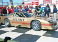 A gold Chevrolet Camaro race car with "Elliott" and "Budweiser" logos is parked on a checkered podium at Laguna Seca during the Rolex Monterey Motorsports Reunion, surrounded by people and cameras at this outdoor IROC event.