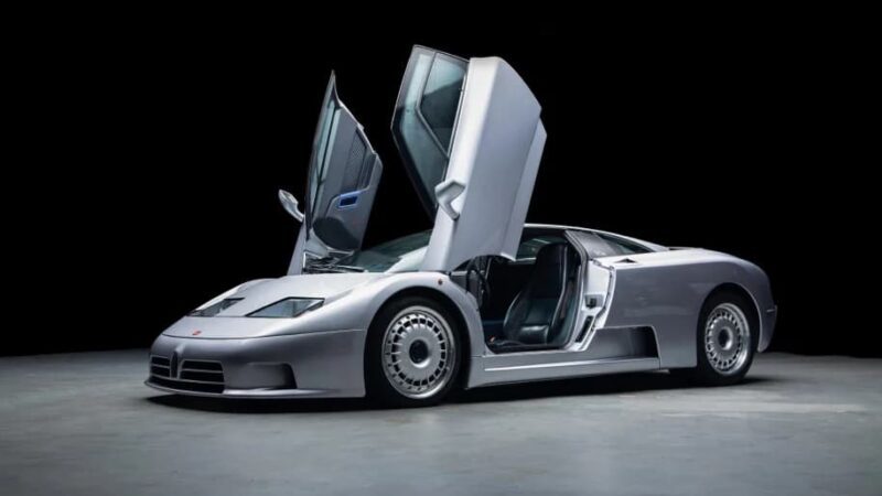 A silver sports car, one of the best manual supercars for sale, with both scissor doors open, parked on a gray surface against a black background.
