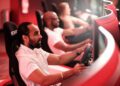 Three people sit in racing simulator chairs at One Porsche Drive in Atlanta, each holding a steering wheel and focused on separate large monitors in a red-lit room.