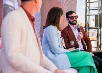 Three people sit on stage during a panel discussion. The man on the right, Enzo Mattioli Ferrari, holds a microphone and speaks, while the other two listen, engaged in the interview about his lasting legacy.
