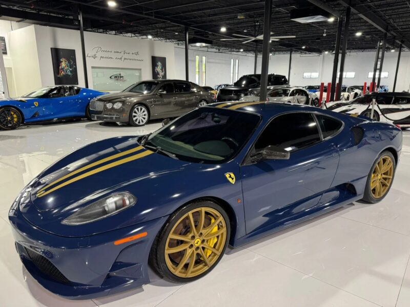 A dark blue Ferrari 430 Scuderia with gold racing stripes and gold wheels, featuring the iconic Prancing Horse, is parked indoors alongside other luxury cars in a showroom, ready for sale.