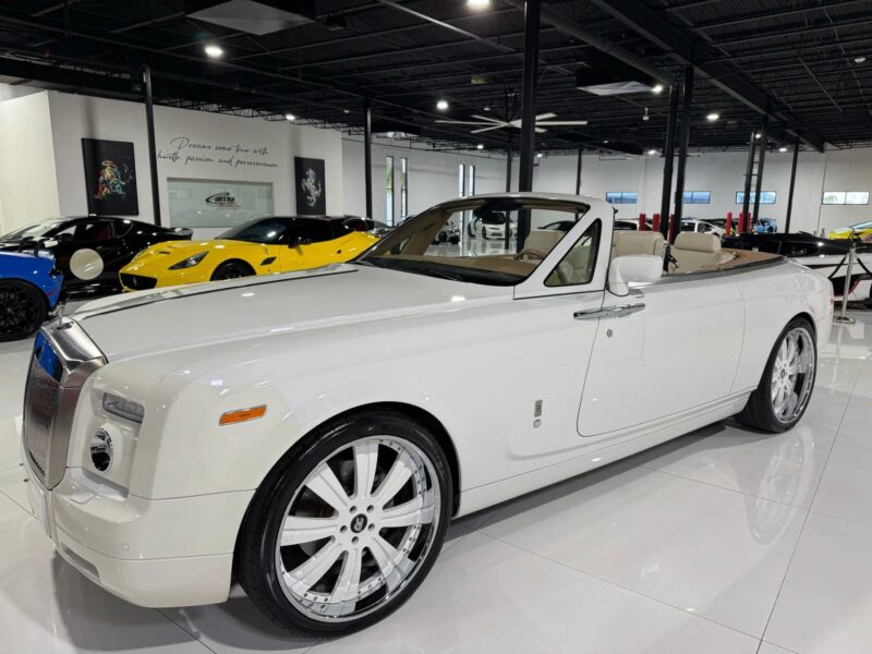 Baltas „Rolls-Royce“ fantominis kabrioletas su dideliais chromo ratais yra pastatytas patalpose salone, apsuptas kitų prabangių sedano modelių fone.