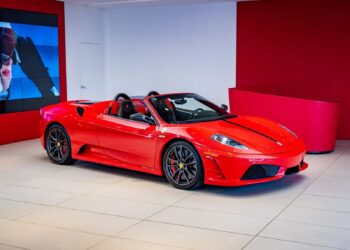 A stunning Ferrari 430 Scuderia, featuring black racing stripes and the iconic Prancing Horse emblem, is displayed in a modern showroom with striking red and white walls and is now for sale.
