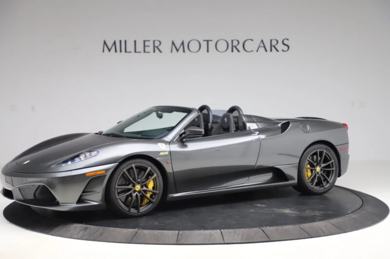 A gray Ferrari 430 Scuderia convertible with black wheels and yellow brake calipers is displayed on a rotating platform in the Miller Motorcars showroom, proudly showcasing the Prancing Horse and available for sale.