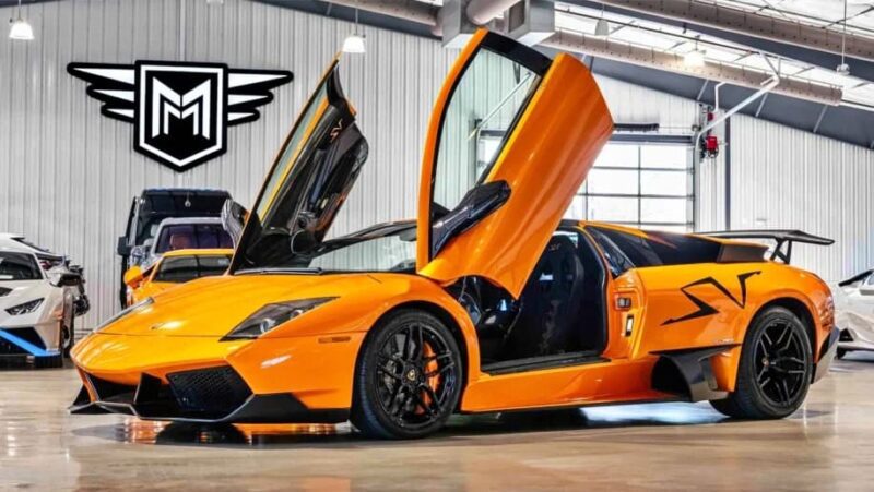 An orange Lamborghini, one of the best manual supercars, with its scissor doors open is parked indoors under bright lighting near a black-and-white logo on the wall.