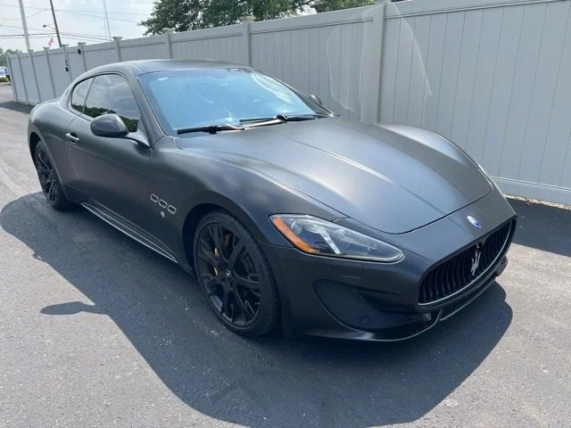A matte black Maserati GranTurismo coupe, the epitome of Italian luxury, is parked on a paved surface next to a white fence under daylight.