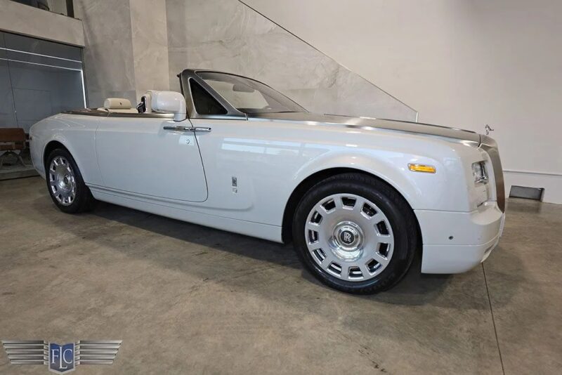 Baltas „Rolls-Royce“ fantominis kabrioletas, prabangaus automobilio pavyzdys, yra pastatytas patalpose ant betoninių grindų prieš paprastą, šviesios spalvos sieną.