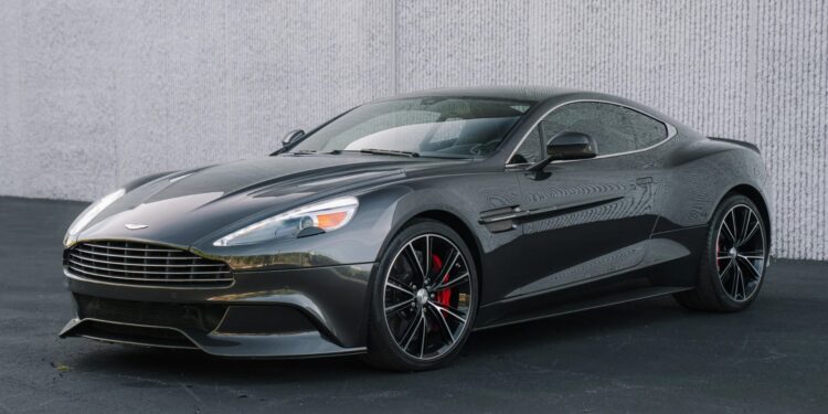 A dark grey Aston Martin sports car, known for its powerful V12 engines, is parked on a blacktop in front of a textured white wall.