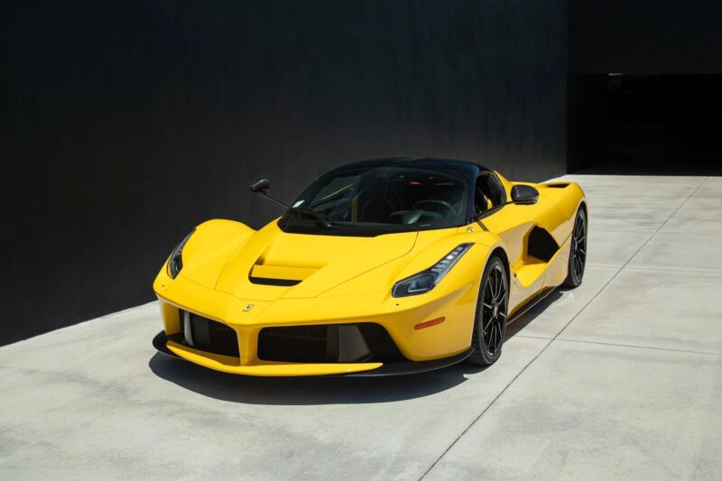 A yellow Ferrari LaFerrari Aperta convertible, showcasing one of the boldest Ferrari colors, is parked on a concrete driveway next to a black building wall.