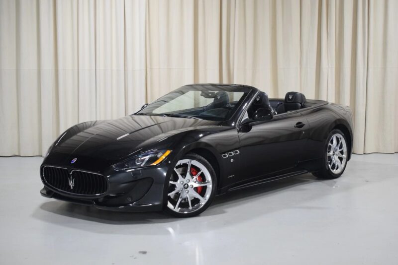 A black Maserati GranTurismo convertible, embodying Italian luxury, sits with the top down on a white floor indoors, positioned elegantly in front of beige curtains.