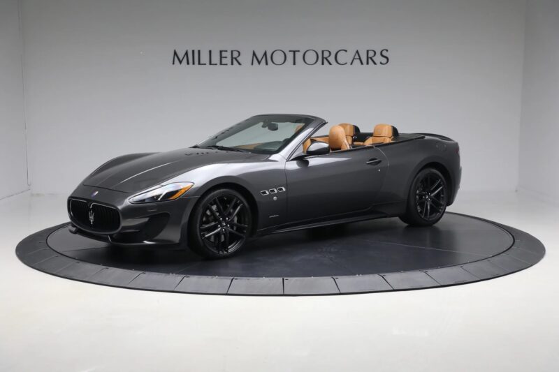 A dark gray Italian luxury Maserati convertible with tan leather interior is displayed on a rotating platform in a showroom labeled "Miller Motorcars.