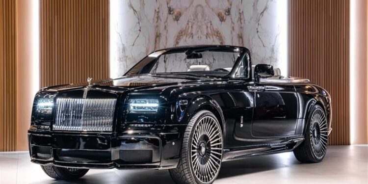 A black Rolls-Royce Phantom convertible, the epitome of a luxury car, is displayed indoors on a light floor with vertical wooden panels and marble walls as the elegant backdrop.