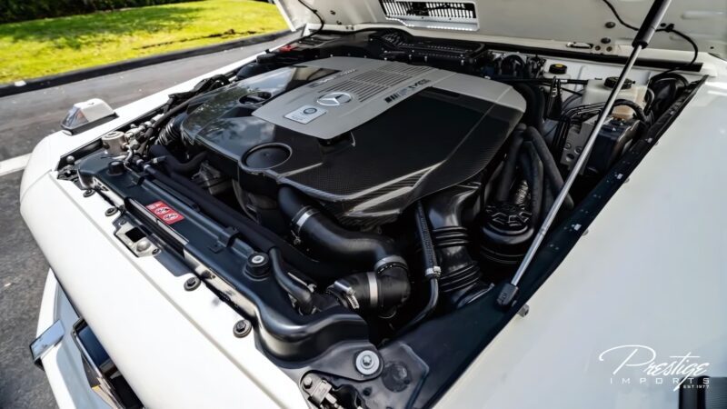 A close-up view of a Mercedes-Benz engine inside the engine bay of a white vehicle, with the hood open and exterior surroundings visible—a rare look under the hood of a Mercedes-Maybach G650 For Sale.