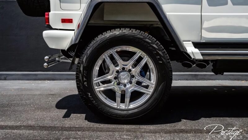 Close-up of a large, chrome-plated wheel and tire on a white Mercedes-Maybach G650 for sale, parked on a paved surface with a black wall in the background.