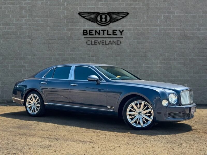 A dark blue Bentley sedan, one of the best Bentleys for sale, is parked in front of a beige brick wall with the Bentley Cleveland logo displayed above it.