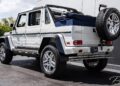 A white Mercedes-Maybach G650 For Sale, this 6x6 SUV boasts large chrome wheels and a rear-mounted spare tire, parked near a building.