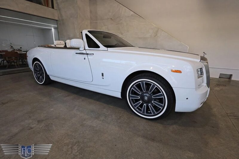 Esminis baltas „Rolls-Royce“ fantominis kabrioletas, pastatytas ant betoninių grindų, su juodais ir sidabriniais lydinio ratais ir įdegio odos interjere, įkūnijant prabangaus sedano eleganciją.