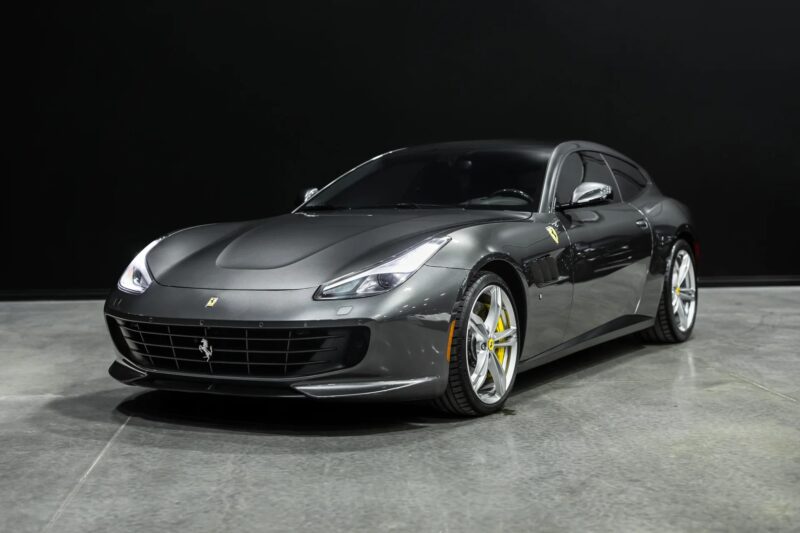 A grey Ferrari sports car with a V12 engine is parked indoors on a concrete floor with a black background.
