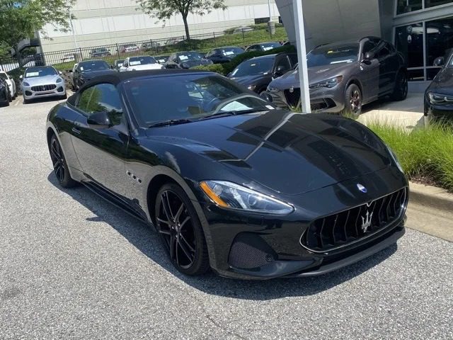 A black Maserati GranTurismo coupe, a pinnacle of Italian luxury, is parked outside a car dealership on a sunny day, surrounded by other vehicles.