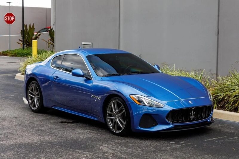 A blue Maserati GranTurismo, a true icon of Italian luxury, is parked on a street near a gray wall, with a stop sign and plants visible in the background.