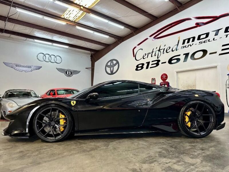 A black Ferrari 488 Pista with yellow brake calipers is parked in an indoor showroom with automotive brand logos on the walls, showcasing this track-ready sports car for sale.