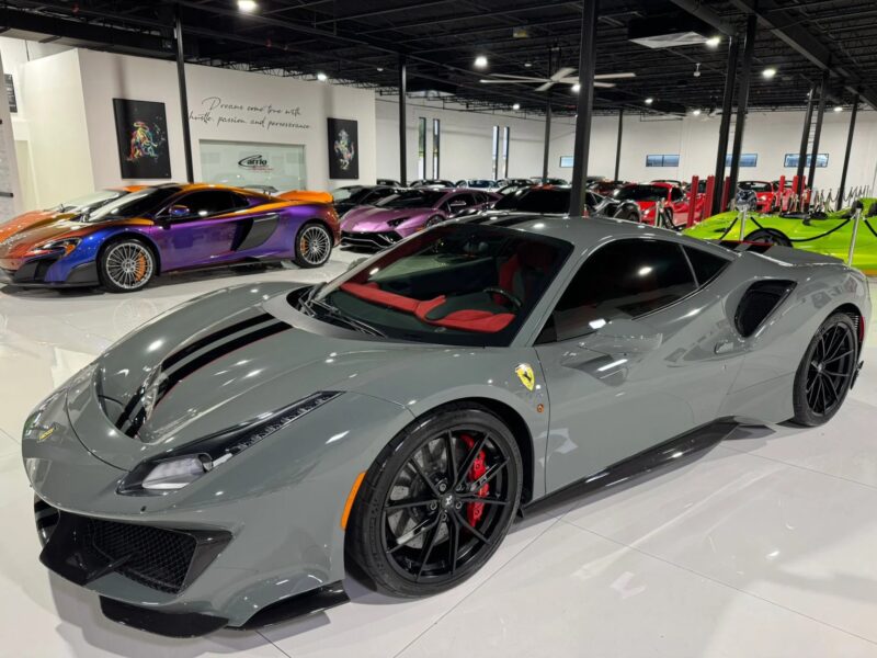A track-ready gray Ferrari 488 Pista with black and red accents is parked indoors, surrounded by other luxury cars.
