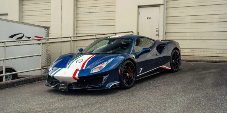 A track-ready blue Ferrari 488 Pista with white and red racing stripes is parked in front of a beige industrial building with closed garage doors.
