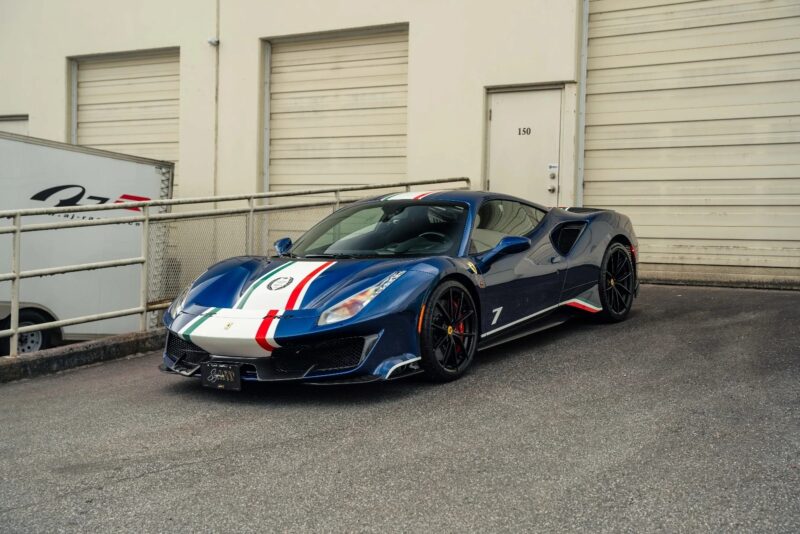 A track-ready blue Ferrari 488 Pista with white and red racing stripes is parked in front of a beige industrial building with closed garage doors.