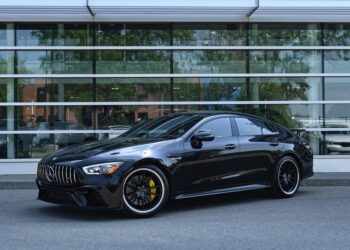 A black Mercedes-Benz AMG luxury car is parked in front of a modern glass building, with reflections visible on the windows behind it.