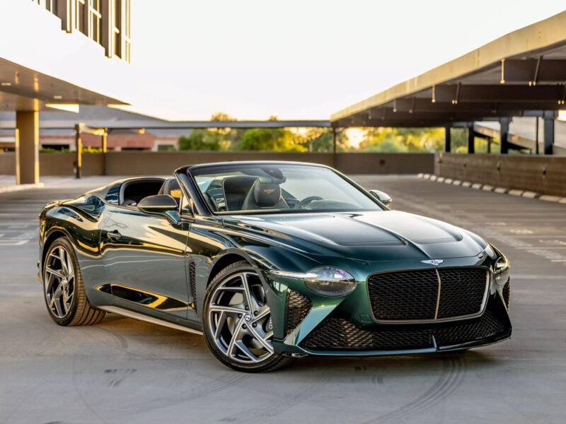 A dark green luxury convertible—one of the best Bentleys—is parked with the top down in an empty, modern parking garage at sunset.