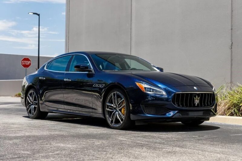 A dark blue Maserati sedan, showcasing Italian luxury, is parked on a paved surface near a gray building and a stop sign under a partly cloudy sky.