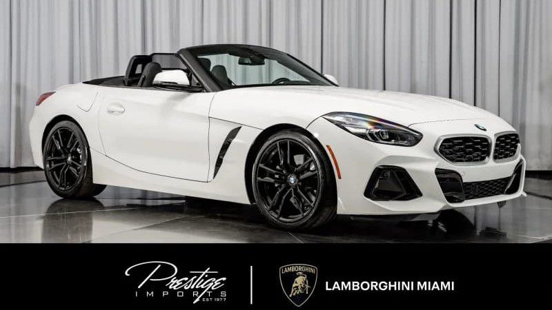 A white BMW Z4 convertible, for sale as one of the best sports cars under $100k, is displayed indoors on a shiny floor with gray curtains in the background. Prestige Imports and Lamborghini Miami logos appear at the bottom.