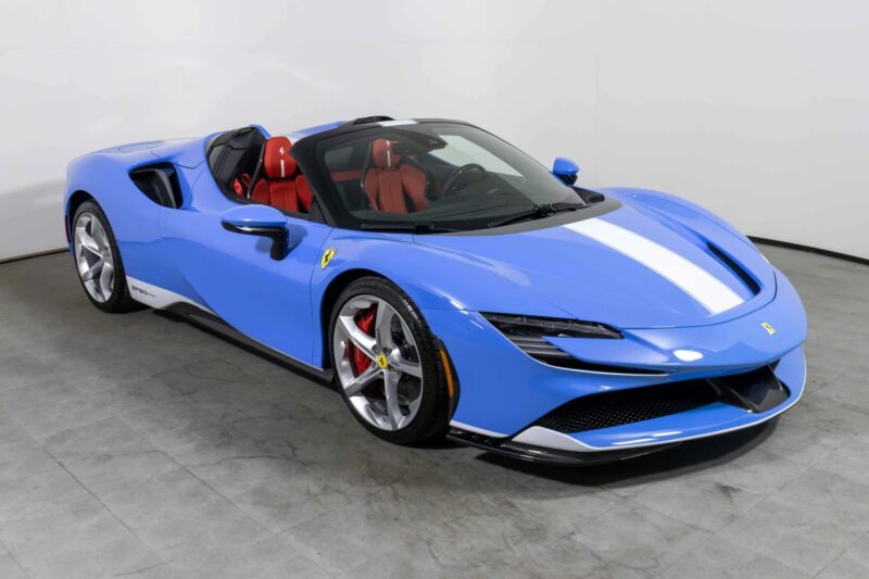A stunning blue Ferrari convertible with a white racing stripe, red interior, and silver wheels showcases classic Ferrari colors as it sits parked indoors on a gray concrete floor.