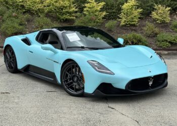 A light blue Maserati sports car, a true example of Italian luxury, is parked on a concrete surface in front of green shrubbery, with a sign visible on the windshield.