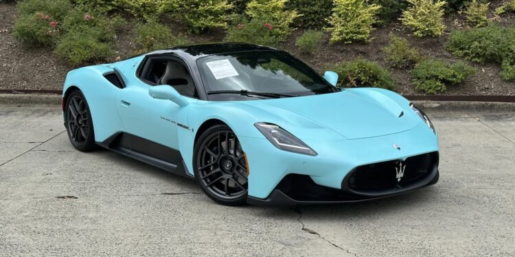 A light blue Maserati sports car, a true example of Italian luxury, is parked on a concrete surface in front of green shrubbery, with a sign visible on the windshield.