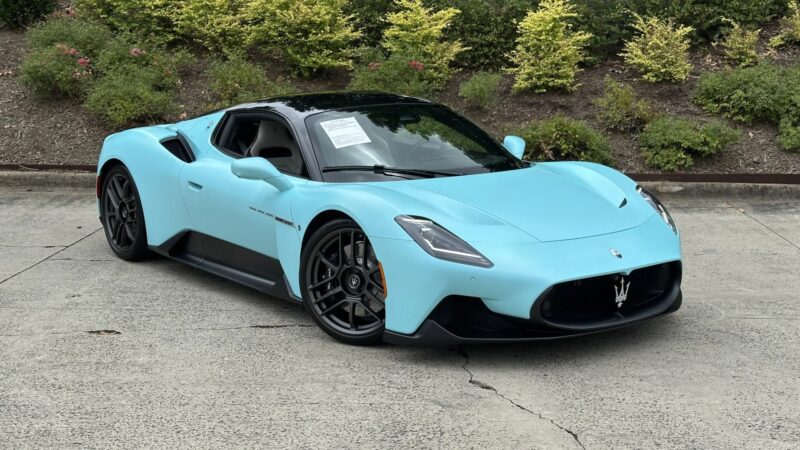 A light blue Maserati sports car, a true example of Italian luxury, is parked on a concrete surface in front of green shrubbery, with a sign visible on the windshield.