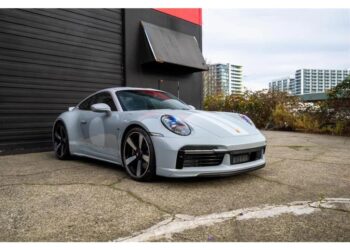 A silver Porsche 911 Sport Classic is parked on a concrete surface beside a dark building, with apartment buildings visible in the background—a striking nod to Porsche history.