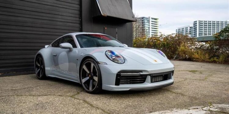 A silver Porsche 911 Sport Classic is parked on a concrete surface beside a dark building, with apartment buildings visible in the background—a striking nod to Porsche history.