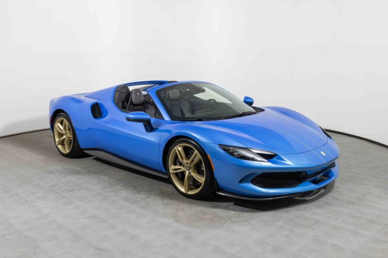 A blue Ferrari convertible sports car with gold wheels is parked indoors on a gray tiled floor against a plain white background, showcasing one of the bold Ferrari colors.