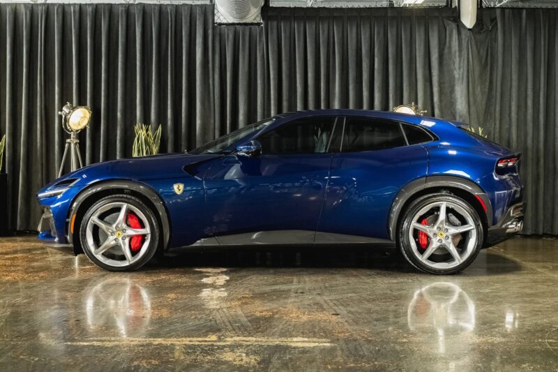 A blue Ferrari Purosangue SUV with red brake calipers is parked indoors on a polished concrete floor in front of black curtains, showcasing why it’s among the best Ferrari SUVs for sale.