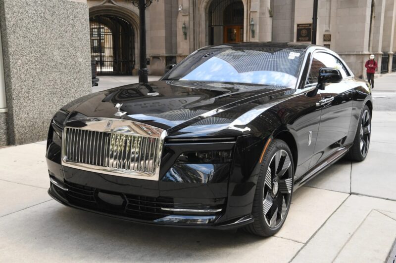 An ultra-luxe black Rolls-Royce Spectre electric super coupe is parked on a city sidewalk near tall buildings.