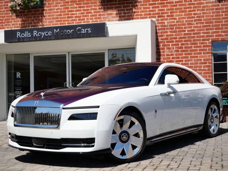 A white and maroon Rolls-Royce Spectre electric super coupe is parked on a cobblestone driveway in front of an ultra-luxe Rolls-Royce Motor Cars dealership with a brick exterior.