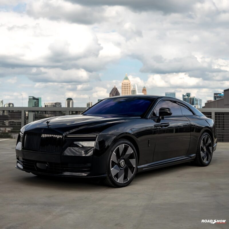 A sleek black Rolls-Royce Spectre, an ultra-luxe electric super coupe, is parked on a rooftop with a city skyline and cloudy sky in the background.