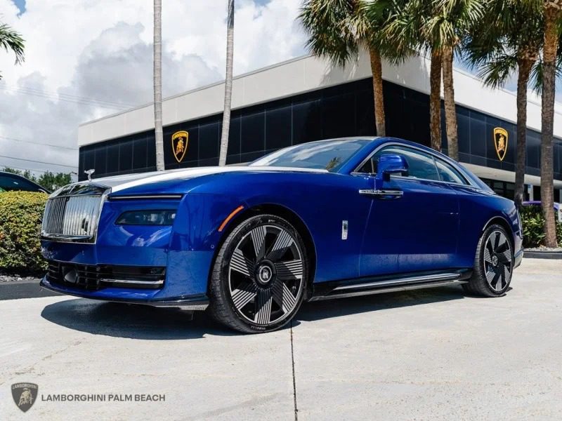 An ultra-luxe blue Rolls-Royce Spectre electric super coupe is parked outside a Lamborghini dealership, framed by palm trees and a modern building in the background.