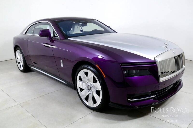 A purple and silver Rolls-Royce Spectre, the ultra-luxe electric super coupe, is parked indoors on a tiled floor, shown from a striking front-side angle.