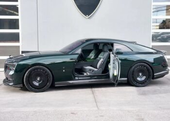 A dark green Rolls-Royce Spectre electric super coupe with its driver-side door open is parked in front of a modern building with large windows.